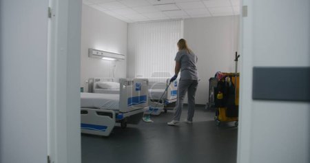 Photo for Adult nurse mops floor between beds in hospital ward. Health worker brings cleaning trolley to hospital room. Female cleaner prepares ward for new patients. Medical staff at work in modern clinic. - Royalty Free Image