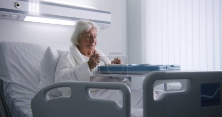 Téléchargez les photos : Femme âgée mange de délicieux aliments et regarde la caméra reposant dans le lit dans la salle d'hôpital lumineux. Patiente âgée se rétablissant après une chirurgie réussie. Installation médicale ou clinique moderne. Portrait. - en image libre de droit