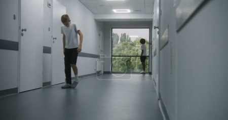 Photo for Injured Caucasian teen with broken leg in bondage walks down medical center corridor to his African American friend in the end of hospital hallway. Boys meet, shake hands and talk near window. - Royalty Free Image