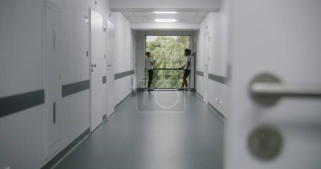 Photo for Caucasian teenager with broken leg stands in the end of medical center hallway, near window. Injured teen talks with his African American friend in hospital corridor. Patient in modern clinic. - Royalty Free Image