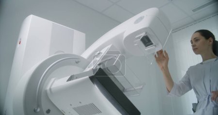 Photo for Hospital radiology room. Female doctor uses computer, then sets up digital mammogram scanning machine for patient before mammography examination. Breast cancer prevention. Modern clinic equipment. - Royalty Free Image