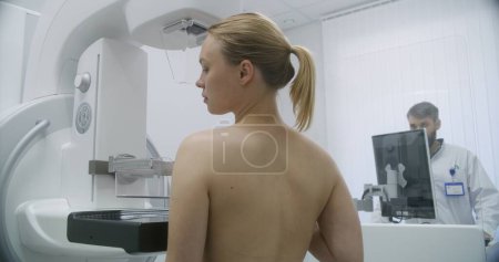 Photo for Happy Caucasian woman stands topless in clinic radiology room. Female patient undergoing mammography scanning examination in modern hospital. Radiologist adjusts mammogram machine using computer. - Royalty Free Image