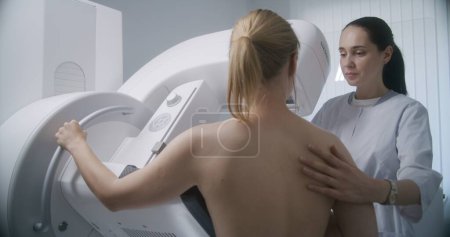 Foto de Sala de radiología del hospital. Mujer caucásica se para durante el procedimiento de detección de mamografía. Médico femenino ajusta mamografía digital para paciente, utiliza ordenador. Prevención del cáncer de mama. Clínica moderna. - Imagen libre de derechos