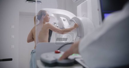 Photo for Adult woman stands topless during mammography screening procedure in hospital radiology room. Radiologist pushes button on control panel to activate digital mammograph using computer. Modern clinic. - Royalty Free Image