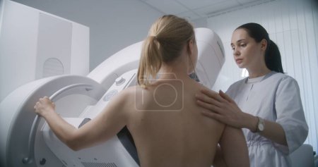 Photo for Hospital radiology room. Caucasian woman stands during mammography screening procedure. Female doctor adjusts digital mammograph for patient, uses computer. Breast cancer prevention. Modern clinic. - Royalty Free Image