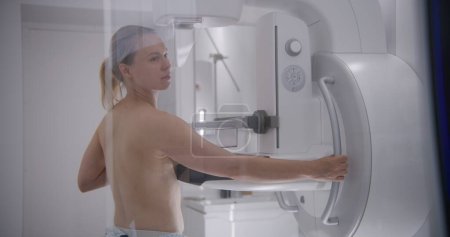 Téléchargez les photos : Femme adulte caucasienne se tient seins nus dans la salle de radiologie de l'hôpital. Patiente soumise à une procédure de dépistage mammographique à l'aide d'un appareil de mammographie. Prévention du cancer du sein. Équipement clinique moderne. - en image libre de droit