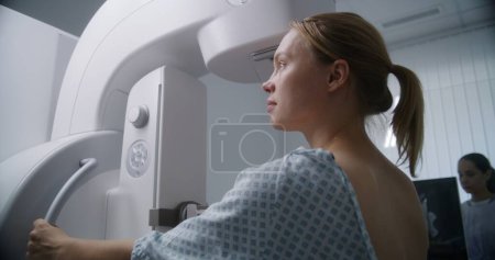 Téléchargez les photos : Femme blanche adulte se tient dans la salle de radiologie de l'hôpital. Patiente soumise à une procédure de dépistage mammographique à l'aide d'un appareil de mammographie numérique. Prévention du cancer du sein. Équipement clinique moderne. - en image libre de droit