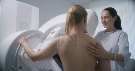 Photo for Hospital radiology room. Caucasian woman stands during mammography screening procedure. Female doctor adjusts digital mammograph for patient, uses computer. Breast cancer prevention. Modern clinic. - Royalty Free Image