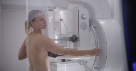 Kaukasische erwachsene Frau steht oben ohne im Radiologie-Raum des Krankenhauses. Patientin, die sich einem Mammographie-Screening mit einem Mammografiegerät unterzieht. Brustkrebsvorbeugung. Moderne Klinik-Ausstattung.
