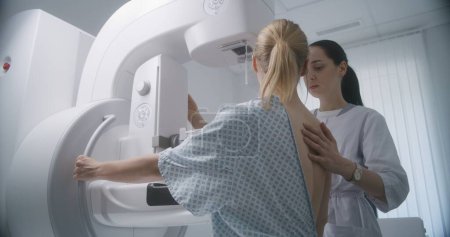 Sala de radiología del hospital. Mujer caucásica se para durante el examen de detección de mamografía en la clínica. El médico femenino ajusta la máquina moderna de la mamografía para el paciente, utiliza la computadora. Prevención del cáncer de mama.
