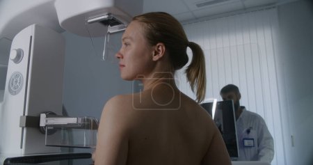 Téléchargez les photos : Femme adulte se tient seins nus subissant un diagnostic de dépistage mammographique dans la salle de radiologie de l'hôpital. Radiologue met en place une machine de mammographie à l'aide d'un ordinateur. Prévention du cancer du sein. Clinique lumineuse moderne. - en image libre de droit