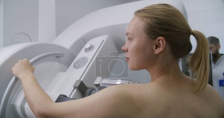 Photo for Adult woman stands topless undergoing mammography scanning checkup in clinic radiology room. Male doctor sets up mammogram machine using computer. Breast cancer prevention. Modern bright hospital. - Royalty Free Image