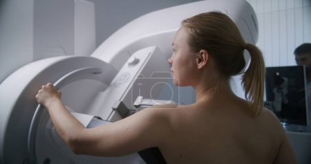 Die erwachsene Frau steht oben ohne zur Mammographie-Untersuchung im Radiologie-Raum der Klinik. Männlicher Arzt richtet Mammografiegerät per Computer ein. Brustkrebsvorbeugung. Modernes, helles Krankenhaus.