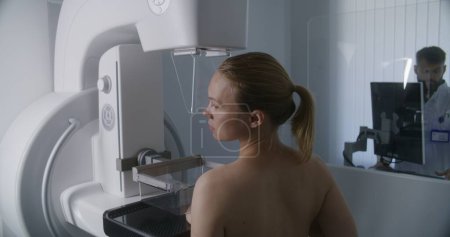 Foto de Mujer caucásica feliz se para en topless en la sala de radiología clínica. Paciente femenina sometida a exploración por mamografía en hospital moderno. Radiólogo ajusta la máquina de mamografía usando computadora. - Imagen libre de derechos