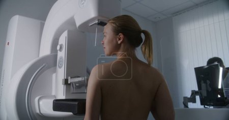 Photo for Adult woman stands topless undergoing mammography screening diagnostic in hospital radiology room. Radiologist sets up mammogram machine using computer. Breast cancer prevention. Modern bright clinic. - Royalty Free Image