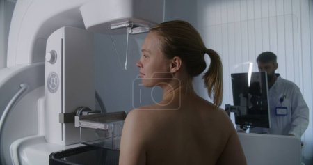 Photo for Adult woman stands topless undergoing mammography screening checkup in clinic radiology room. Male doctor adjusts mammogram machine using computer. Breast cancer prevention. Modern bright hospital. - Royalty Free Image