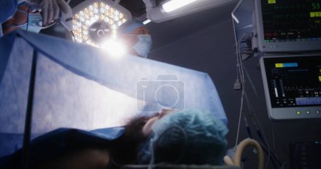 Photo for Adult professional surgeons perform laparoscopy operation and look at monitors in well-equipped surgery room. Nurse assists doctors. Patient lies on table with breathing tube under anesthesia. - Royalty Free Image