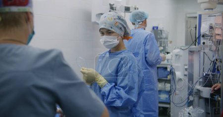 Photo for Professional male surgeon and nurses stand in operating room after long operation. Female medics clean workspace after surgery. Doctor takes off gloves. Medical staff at work in modern clinic. - Royalty Free Image