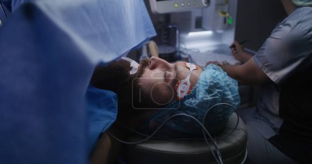 Foto de Primer plano del paciente acostado en la mesa quirúrgica bajo anestesia con tubo respiratorio durante la cirugía. La enfermera mira los monitores, escribe los resultados de la electrocardiografía. Personal médico en el trabajo en quirófano. - Imagen libre de derechos
