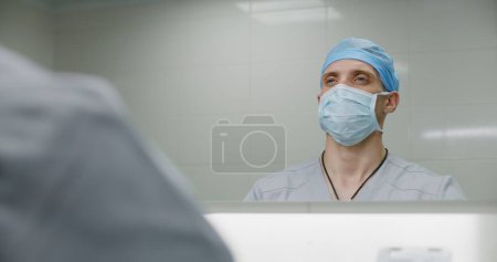 Téléchargez les photos : Chirurgien professionnel en uniforme nettoie les mains avant la chirurgie. Homme médecin se prépare à effectuer une opération chirurgicale avec patient gravement blessé. Le personnel médical travaille dans un hôpital moderne. Gros plan. - en image libre de droit