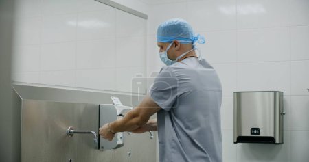 Foto de Cirujano profesional en uniforme se lava las manos antes de la operación quirúrgica. El médico o paramédico se prepara para realizar una cirugía con un paciente gravemente lesionado. El personal trabaja en instalaciones médicas modernas. - Imagen libre de derechos