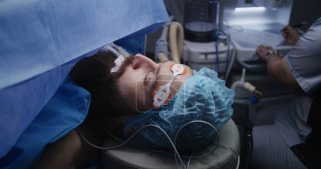 Photo for Close up of patient lying on surgical table under anesthesia with breathing tube during surgery. Nurse looks at monitors, writes electrocardiography results. Medical staff at work in operating room. - Royalty Free Image