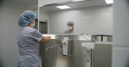 Photo for Surgeon in uniform washes hands with soap before surgery. Female medic prepares to performing surgical operation with seriously injured patient. Medical personnel work in modern hospital. Back view. - Royalty Free Image