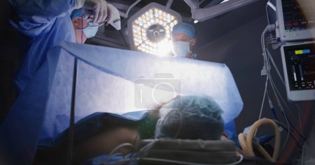 Photo for Adult professional surgeons perform laparoscopy operation and look at monitors in well-equipped surgery room. Nurse assists doctors. Patient lies on table with breathing tube under anesthesia. - Royalty Free Image