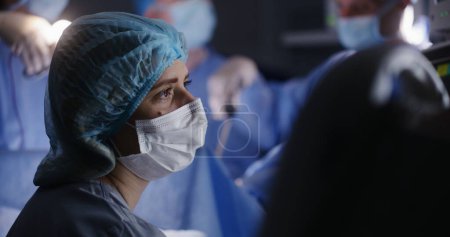 Téléchargez les photos : Le médecin féminin se trouve dans la salle d'opération moderne et vérifie l'état du patient sur les moniteurs d'électrocardiographie. Chirurgiens professionnels masculins en costume chirurgical effectuer une chirurgie à l'aide d'instruments de laparoscopie. - en image libre de droit
