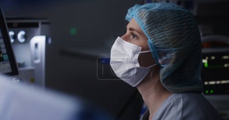 Téléchargez les photos : Femme adulte médecin en chapeau médical et masque se trouve dans la salle d'opération moderne et parle avec les chirurgiens. L'infirmière examine le moniteur d'électrocardiographie et vérifie l'état du patient pendant une chirurgie difficile. - en image libre de droit
