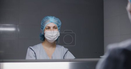 Photo for Surgeon in uniform cleans hands before surgery and looks at mirror. Female nurse prepares to performing surgical operating with seriously injured patient. Medic works in medical facility. Back view. - Royalty Free Image
