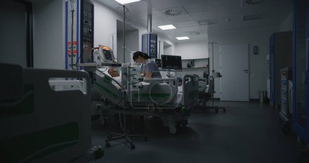 Téléchargez les photos : Salle d'urgence avec équipement moderne à l'hôpital. Homme âgé dans le masque à oxygène se trouve et dort au lit après une chirurgie réussie. Infirmière travaille sur ordinateur dans le département de soins intensifs de l'établissement médical. - en image libre de droit