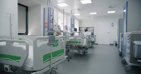 Emergency room with modern equipment in hospital. Elderly man in oxygen mask sleeps in bed after successful surgery. Nurse takes care of old patient. Intensive care department in medical facility.