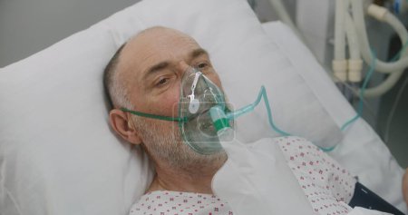 Téléchargez les photos : L'infirmière s'occupe du patient âgé malade. Homme âgé dans le masque à oxygène se trouve dans le lit pendant la ventilation pulmonaire artificielle. Urgences de l'hôpital moderne. Service de coronavirus en soins intensifs en clinique. - en image libre de droit