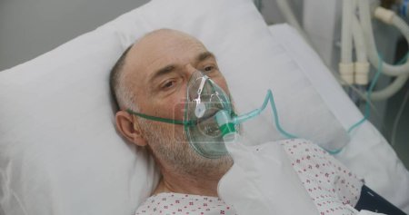 Photo for Nurse takes care of sick senior patient. Elderly man in oxygen mask lies in bed during artificial lung ventilation. Emergency room in modern hospital. Intensive care coronavirus department in clinic. - Royalty Free Image