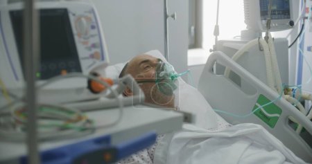 Sick elderly man in oxygen mask in bed in hospital ward. Old patient with pneumonia during lung ventilation. Modern emergency room in clinic. Intensive care coronavirus department in medical facility.