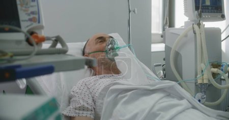 Foto de Hombre anciano enfermo con máscara de oxígeno en la cama en el hospital. Paciente anciano con neumonía durante la ventilación pulmonar. Moderna sala de emergencias en clínica. Departamento de coronavirus de cuidados intensivos en centros médicos. - Imagen libre de derechos