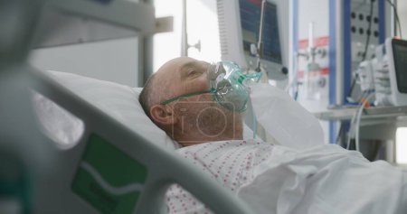 Téléchargez les photos : Homme âgé dans le masque à oxygène se trouve dans le lit pendant la ventilation pulmonaire artificielle. L'infirmière s'occupe du patient. Urgences dans une clinique moderne. Service de coronavirus de soins intensifs dans un établissement médical. - en image libre de droit