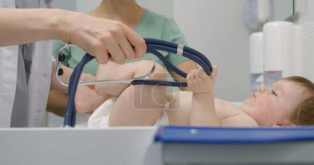 Photo for Close up of female doctor checking development of hip joints of baby. Little boy lies on changing table in light hospital ward. Mother stands and holds her child on appointment with pediatrician. - Royalty Free Image