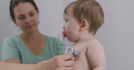 Photo for Handsome child sits at doctor checkup and puts pacifier in mouth. Pediatrician uses stethoscope to listen heartbeat and lungs of little kid. Mother with cute baby in hospital. Close up. Slow motion. - Royalty Free Image
