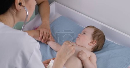 Photo for Mother holds hand of cute baby with pacifier sitting on changing table. Doctor uses stethoscope to listen heartbeat and lungs of little kid. Pediatrician does checkup in clinic. Close up. Slow motion. - Royalty Free Image