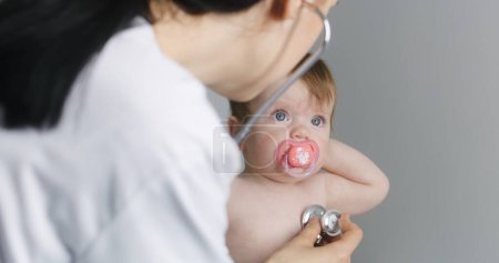 Photo for Cute baby sits on changing table, takes out pacifier from mouth, waves hands, cries. Female pediatrician tries to listen heartbeat of kid using stethoscope. Mom with little child on checkup. Close up. - Royalty Free Image