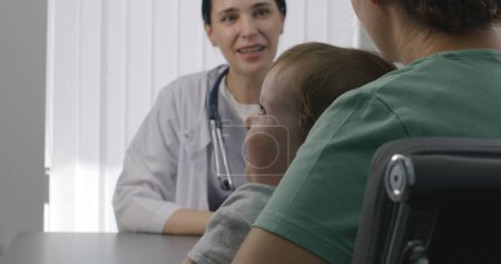 Téléchargez les photos : Une pédiatre consulte une patiente avec un mignon petit enfant sur rendez-vous à l'hôpital. La femme est assise sur la chaise au bilan médical. Médecin amical travaille dans le centre médical moderne. Ferme là. Vue arrière. - en image libre de droit