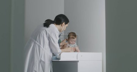 Foto de Amable pediatra mujer da juguete a bebé guapo sentado en el cambiador. El médico usa el estetoscopio para escuchar los latidos del corazón y los pulmones de un niño pequeño y lindo. El médico hace chequeo en el hospital. - Imagen libre de derechos