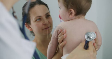 Photo for Happy mother holds on hands her handsome baby with pacifier in mouth. Pediatrician uses stethoscope to listen to heartbeat and lungs of cute little child. Physician does checkup in hospital. Close up. - Royalty Free Image