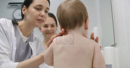 Photo for Little boy sits on changing table on appointment with pediatrician in light hospital ward. Mother holds her baby. Female doctor uses stethoscope and checks condition of kid. Medic at work in clinic. - Royalty Free Image