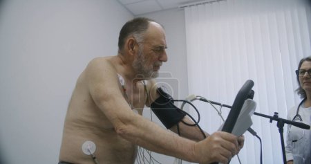 Téléchargez les photos : Homme âgé avec capteurs holter pédales orbitrek dans la salle de cardiologie. Une cardiologue diagnostique la guérison d'un patient atteint d'une maladie cardiovasculaire. Réhabilitation fonctionnelle dans un hôpital moderne. - en image libre de droit