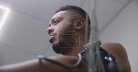 Photo for African American man pedals orbitrek in cardiology room during functional rehabilitation. Adult patient with heart or cardiovascular disease. Electrocardiography in modern clinic or hospital. Portrait - Royalty Free Image