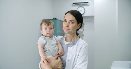 Photo for Female pediatrician stands in middle of bright hospital room and looks at camera. Adult doctors holds baby. Little boy looks at camera and holds hand of medic. Medical staff at work in modern clinic. - Royalty Free Image