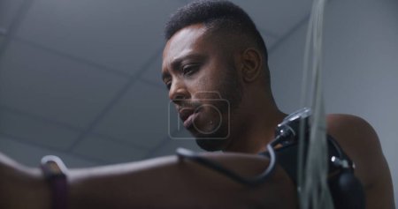 Photo for African American man with tonometer and sensors pedals orbitrek in cardiology room during holter monitoring procedure. Patient with cardiovascular disease. Electrocardiography in hospital. Portrait. - Royalty Free Image
